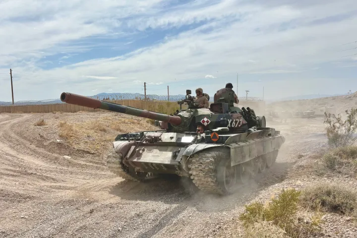 Military tank on dusty road with three soldiers, desert terrain, clear sky.