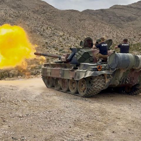 Tank firing with large muzzle flash in a rocky desert, staff seated on turret.