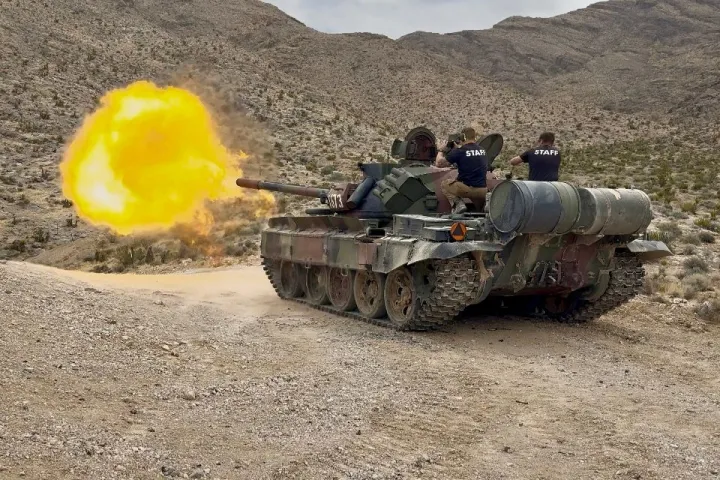 Tank firing with large muzzle flash in a rocky desert, staff seated on turret.